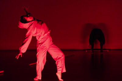 dans une lumière baignée de couleur rouge, le personnage du petit chaperon rouge, debout sur la pointe des pieds, visage en arrière sous une capuche, agite ses bras tendus vers l'arrière de son corps. en fond de scène, la silhouette du Loup, tête penchée en avant vers le sol dans une position d'attente inquiétante - Agrandir l'image 8 sur 10, fenêtre modale