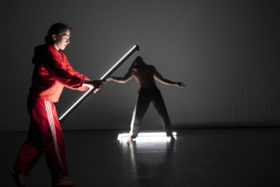 sur scène au premier plan à gauche de l'image se tient de profil debout la danseuse en survêtement rouge; tenant à bout de bras une barre métallique, tandis qu'en arrière-plan, la silhouette de 'linterprète du loup semble par illusion d'optique recevoir un coup de cette barre sur la tête, bras écartés de surprise et de douleur - Agrandir l'image 9 sur 10, fenêtre modale