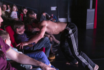 Torse nu en pantalon de survêtement noir à bandes blanches, le danseur qui interprète le loup apparaît debout, de profil, dans les rangées où se tient le public, il mime le geste de se jeter sur un garçonnet assis au premier rang - Agrandir l'image 6 sur 10, fenêtre modale