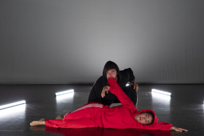 allongé sur le côté face au public, l'interprète du petit chaperon rouge, en jogging à capuche rouge, pieds nus et visage exprimant le dépit, est saisie par la manche entre les mâchoires du loup, en jogging à capuche noir agenouillé face public - Agrandir l'image 4 sur 10, fenêtre modale