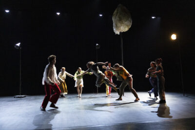 Sur scène, le sol éclairé par une lumière argentée, un groupe de comédiens en mouvement sont rassemblés en un cercle où chaque individualité exprime sa propre gestuelle. En fond de scène les lumières ténues de projecteurs - Agrandir l'image 2 sur 6, fenêtre modale