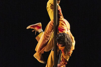 Plan rapproché sur une circassienne aux cheveux bruns mi-longs à trois jambes en baskets, vêtue d'une combinaison ample de couleurs chaudes, pendue tête en bas par les pieds à un mât chinois - Agrandir l'image 3 sur 6, fenêtre modale