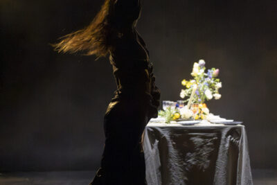 Sur les planches de l'Escale de Tournefeuille, une jeune femme debout en ombres chinoises, se tient debout et cambrée, bras levés en l'air, aux abords d'une table nappée de blanc aux broderies en dentelle traditionnelle, un vase fleuri disposé dessus. - Agrandir l'image 4 sur 6, fenêtre modale