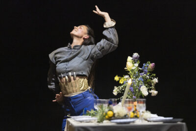une comédienne, ceinturée d'une rangée fantaisiste de couverts de table, dans une posture de flamenco se tient menton levé, bras et mains ourlés en une pose artistique derrière une table joliment agrémentée de fleurs des champs, le tout composant une image très esthétique - Agrandir l'image 5 sur 6, fenêtre modale