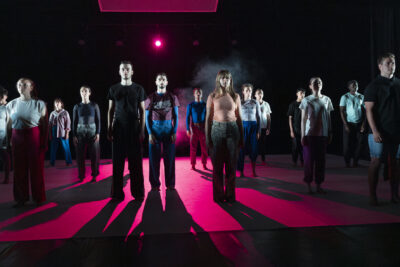 Debout sur scène, alignés face public, en tenue décontractée tee-shirt et jogging, les danseurs se tiennent bien droits dans les rais de lumière rose fushia des projecteurs - Agrandir l'image 1 sur 6, fenêtre modale