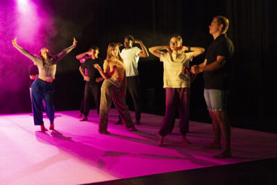 Dans la lumière rose fushia d'un projecteur de scène, une partie du groupe des danseurs se tient alignée, bras écartés à hauteur de leur cou, tandis qu'en arrière-plan, à gauche de l'image une jeune fille en position déhanchée, lève les bras au ciel en un mouvement libérateur - Agrandir l'image 3 sur 6, fenêtre modale