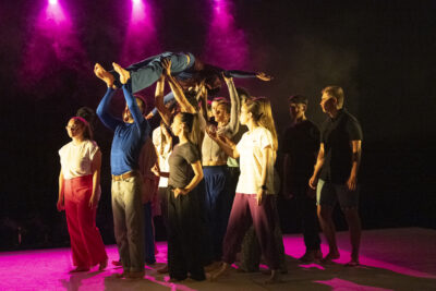 Dans les lumières de scène rose fushia, de jeunes danseurs de profil, debout, tiennent à bout de bras un corps allongé et figé au-dessus de leurs têtes - Agrandir l'image 2 sur 6, fenêtre modale