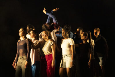 seul élément éclairé dans la pénombre sur scène, un groupe compact de danseurs debout se tient de façon solennelle dans la lumière blanche, un danseur se trouve en hauteur au-dessus de leurs têtes, allongé, les bras en mouvement au-dessus de la mêlée - Agrandir l'image 4 sur 6, fenêtre modale