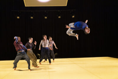 Sur la scène éclairée, au-dessus du plancher couleur crème, un danseur effectue un salto arrière impressionnant, corps entièrement renversé à l'horizontale à 1 ou 2 m de hauteur du sol, tandis qu'un petit groupe de danseurs en mouvement se trouve debout à droite de l'image en train de l'observer d'un air interdit - Agrandir l'image 5 sur 6, fenêtre modale
