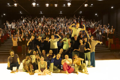 Dans cette image de fin de spectacle, les nombreux des interprètes de la pièce se tiennent réunis en souriant et effectuant des gestes enthousiastes, en une photo joyeuse de groupe face caméra et dos au public debout en train d'applaudir sous les lumières rallumées de la salle - Agrandir l'image 6 sur 6, fenêtre modale