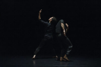 Sur scène, dans le noir où seuls affleurent les couleurs de leur peau, dans une posture de lâcher-prise et tendue à la fois, le couple de danseurs se tient mutuellement dans un mouvement giratoire debout - Agrandir l'image 2 sur 4, fenêtre modale