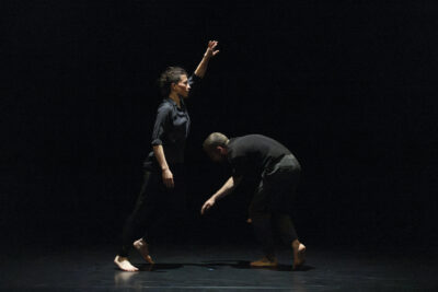 De profil, à gauche la danseuse en pantalon et chemise noire pieds nus se tient délicatement sur un pied, le reste du corps tendu vers l'avant et vers le haut, tandis qu'à droite son comparse masculin vêtu de même, boucle le mouvement circulaire que forment leurs deux corps dans une position d'agenouillement;, bras tendu vers le sol comme en train de cueillir quelque chose - Agrandir l'image 3 sur 4, fenêtre modale