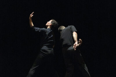 dans une posture très artistique se tiennent les deux danseurs virevoltant debout dans le noir, leurs têtes enlacées l'une à l'autre, elle levant un bras gauche à hauteur de son visage, lui de dos face à la caméra balançant son bras droit vers l'arrière dans un mouvement harmonieux de leurs deux corps coordonné - Agrandir l'image 4 sur 4, fenêtre modale