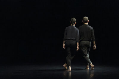 Fond noir, sol aux reflets sombres et mats, pénombre, deux danseurs debout, de dos, cheveux plaqués, en tenue sobre, se tiennent côte à côte en se fondant dans cet espace - Agrandir l'image 1 sur 4, fenêtre modale