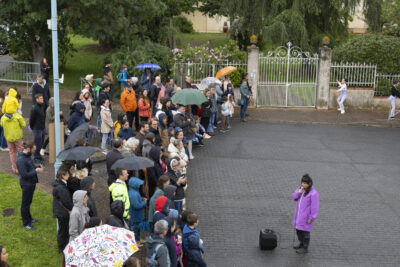 Dans la rue, devant l'Ecole de danse de la Ville de Tournefeuille, une comédienne vêtue d'une parka violette s'adresse au public par le biais d'un micro connecté à une enceinte, tous l'écoutent attentivement. - Agrandir l'image 2 sur 6, fenêtre modale