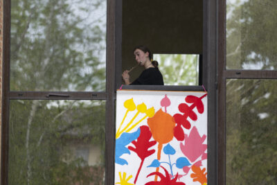 À la fenêtre de L'EEA on aperçoist une jeune fille en train de jouer de la clarinette. Les baies vitrées reflètent une nature luxuriante. Sous la fenêtre une affiche représente de manière colorée des feuilles d'arbres. - Agrandir l'image 6 sur 6, fenêtre modale