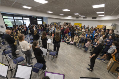 Un orchestre se tient devant un large public de tout âges on y aperçois la cheffe d'orchestre en noir au centre. Tous sont abrités dans l'Ecole d'Enseignements Artistiques de Tournefeuille. - Agrandir l'image 1 sur 6, fenêtre modale
