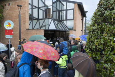 Un large public de tous âges arborant des parapluies s'amoncelle devant l'entrée de l'EEA. On aperçoit au loin quelques musiciens devant leurs pupitres. - Agrandir l'image 5 sur 6, fenêtre modale