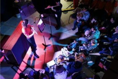 vue d'ensemble en plongée, sur un parterre de public assis autour d'une flûtiste sur scène dans une jolie lumière rose-violette