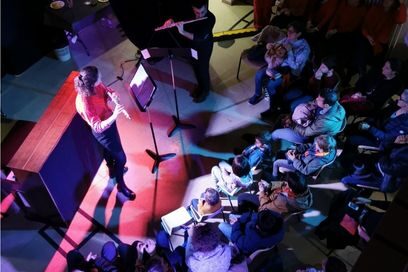 vue d'ensemble en plongée, sur un parterre de public assis autour d'une flûtiste sur scène dans une jolie lumière rose-violette - Agrandir l'image, fenêtre modale