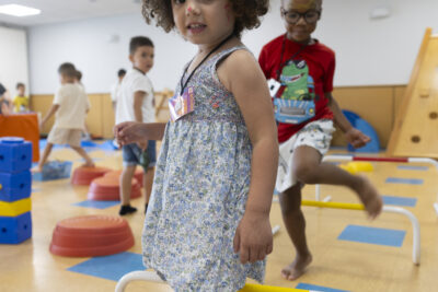 Photo d'une petite fille qui sourit à l'objectif tandis qu'elle enjambe une barre au sol qui fait partie d'un parcours plus large, on voit un autre enfant derrière elle - Agrandir l'image 5 sur 15, fenêtre modale