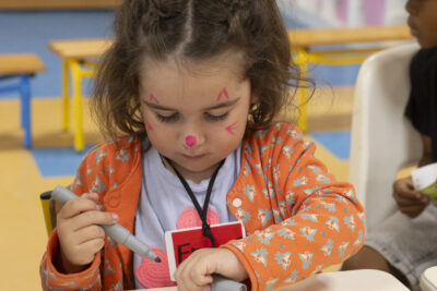 Une petite fille avec des motifs roses sur le visage dessine sur son support papier avec concentration - Agrandir l'image 6 sur 15, fenêtre modale
