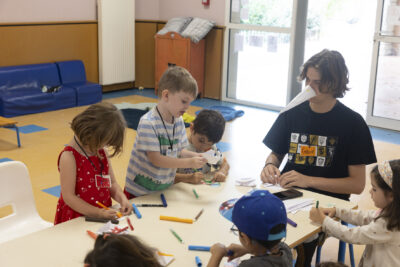 Photo d'enfants en train de dessiner et plier des papiers autour d'une table, un animateur porte un bec d'oiseau en papier - Agrandir l'image 7 sur 15, fenêtre modale