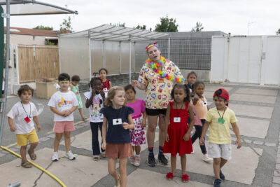 Une dizaine d'enfants réunis dans une cour avec une animatrice qui porte un collier à fleurs - Agrandir l'image 8 sur 15, fenêtre modale
