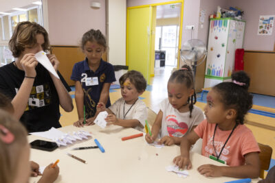 Photo d'enfants en train de dessiner et plier des papiers autour d'une table, un animateur porte un bec d'oiseau en papier - Agrandir l'image 12 sur 15, fenêtre modale