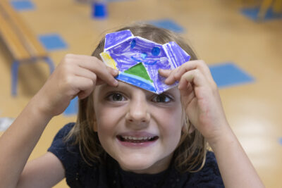 Une enfant en gros plan sourit à l'objectif en montrant un dessin qu'elle tient sur son front - Agrandir l'image 1 sur 15, fenêtre modale