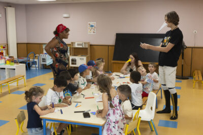 Photo d'une douzaine d'enfants rassemblés autour d'une table en train de dessiner, deux animateurs - Agrandir l'image 15 sur 15, fenêtre modale