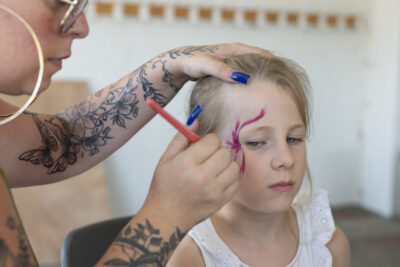 Photo d'une jeune fille blonde qui se fait maquiller un motif de fleur sur le visage par une animatrice - Agrandir l'image 10 sur 18, fenêtre modale
