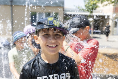 Photo d'un jeune garçon sous des jets d'eau, des gouttelettes autour de lui, la casquette et le tee-shirt trempés - Agrandir l'image 1 sur 18, fenêtre modale