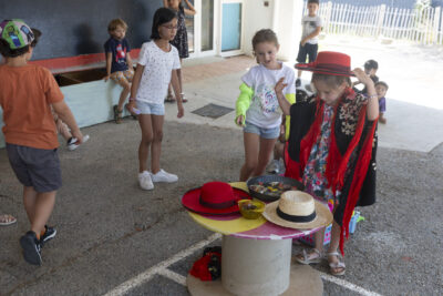 Photo d'enfants sous un préau, des jeunes filles autour d'une petite table ronde sur laquelle il y a des chapeaux à l'espagnole/flamenco, une en essaie un avec une sorte de châle noir et rouge - Agrandir l'image 8 sur 18, fenêtre modale