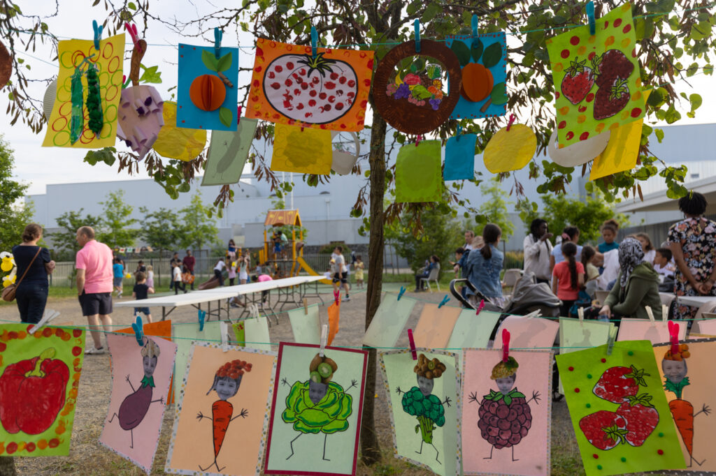 Photo de dessins d'enfants accrochés et derrière on voit, sur une place en extérieur, des tables et des gens - Agrandir l'image, fenêtre modale