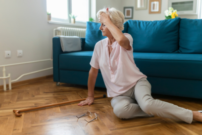 Photo d'une femme âgée dans un salon, assise par terre après avoir chuté, sa canne est au sol et elle a une main sur le front - Agrandir l'image, fenêtre modale
