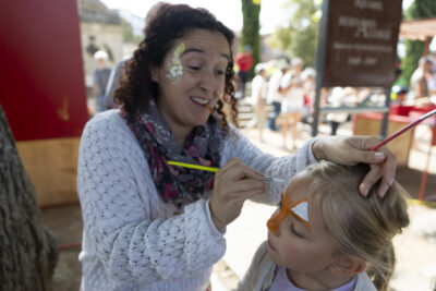 Photo d'une jeune femme qui maquille une jeune fille en extérieur, l'arrière-plan est flou mais on aperçoit des gens - Agrandir l'image 6 sur 22, fenêtre modale