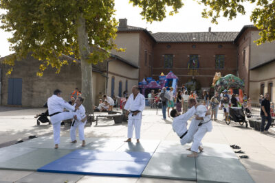 Photo d'une démonstration de sport de kimino, probablement du judo, sur des tatamis sur le parvis de la Mairie. On voit à l'arrière-plan des jeux gonflables géants et la mairie - Agrandir l'image 3 sur 22, fenêtre modale