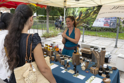 Photo d'un stand d'onguents avec une femme derrière le stand qui échange avec deux jeunes femmes qu'on voit de dos devant le stand - Agrandir l'image 11 sur 22, fenêtre modale