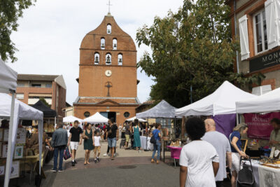 photo paysage prise depuis l'impasse Baylac, avec vue sur l'église au fond et des stands de chaque côté de la rue, des gens qui vont de l'un à l'autre - Agrandir l'image 15 sur 22, fenêtre modale