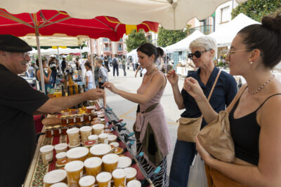 photo paysage d'un stand de vente de miel, trois femmes sont devant et l'homme qui tient le stand fait goûter du miel - Agrandir l'image 17 sur 22, fenêtre modale