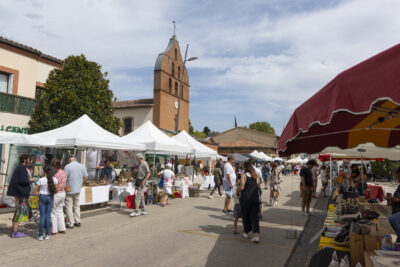 Photo paysage de la rue Doumergue avec vue sur l'église, et sur les stands des producteurs et commerçants de chaque côté de la rue piétonnisée. Des personnes se baladent de stand en stand, la journée est ensoleillée - Agrandir l'image 18 sur 22, fenêtre modale