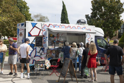 photo paysage d'un foodtruck blanc avec des formes bleues qui évoquent la Grèce, devant lequel une dizaine de personnes font la queue - Agrandir l'image 19 sur 22, fenêtre modale