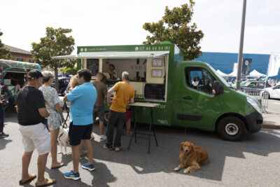 Photo paysage d'un foodtruck vert ouvert devant lequel 5 ou 6 personnes font la queue. Un chien est aussi allongé à l'ombre à côté des gens. - Agrandir l'image 20 sur 22, fenêtre modale