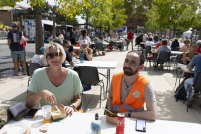 Photo paysage d'une femme et d'un homme, lui porte un gilet orange de l'organisateur Made in Tounefeuille, à table en train de manger sur l'allée devant la Mairie. Derrière eux, à table également, de nombreuses personnes, alors qu'il fait beau temps - Agrandir l'image 21 sur 22, fenêtre modale
