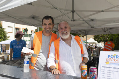 Photo paysage de deux hommes bénévoles portant le gilet orange avec le logo de Made in Tournefeuille, en train de tenir la buvette, tout sourire à l'objectif - Agrandir l'image 22 sur 22, fenêtre modale
