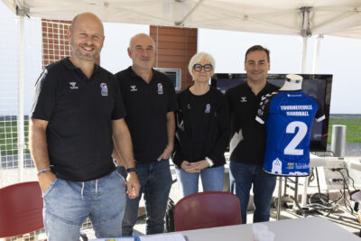 photo d'un stand associatif, trois hommes et une femme en ligne derrière le stand sourient à l'objectif, ils portent un polo noir avec le logo THB, et à côté d'eux un mannequin porte un maillot sportif bleu sur lequel il est écrit "Tournefeuille handball" - Agrandir l'image 8 sur 68, fenêtre modale