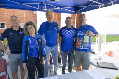 photo d'un stand associatif, quatre homme et une femme derrière le stand, avec un haut bleu, sourient à l'objectif, l'un d'eux tient sur lui une affiche sur le 10 km et semi-marathon de Tournefeuille qu'il montre - Agrandir l'image 9 sur 68, fenêtre modale