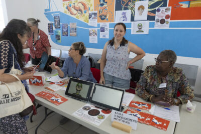 photo du stand de l'école d'enseignements artistiques avec trois femmes derrière, une au milieu est debout et sourit à l'objectif, une autre femme est devant le stand et regarde les plaquettes informatives - Agrandir l'image 13 sur 68, fenêtre modale