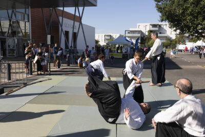 Photo d'une démonstration d'aïkido sur tatamis, en extérieur sur le parking du Phare. On voit quelques familles regarder et des stands associatifs à l'arrière-plan - Agrandir l'image 2 sur 68, fenêtre modale